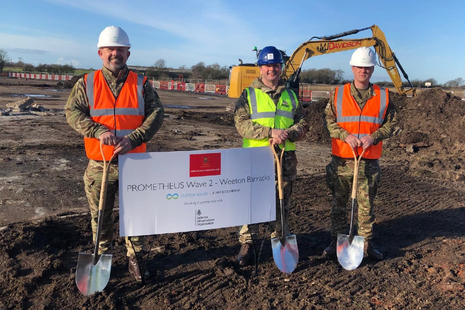 Three men in Army uniforms, high vis vests and construction helmets stand on a large area of soil holding spades. Behind them is a digger and they hold a sign between them reading 'Prometheus Wave 2 - Weeton Barracks'.