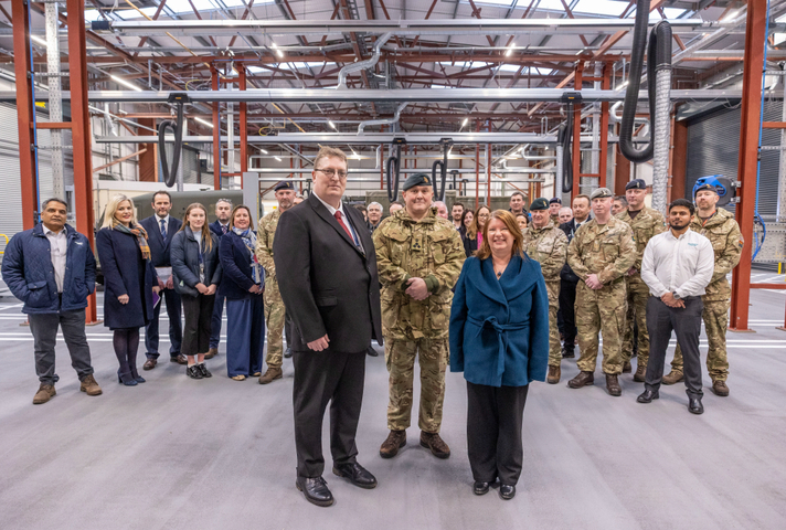 A large group of people, some wearing civilian dress and others in Army uniform, stand facing the camera in a large industrial building. Three are at the front, a man in a suit, a woman in a blue coat, and a man in a Brigadier's uniform. 