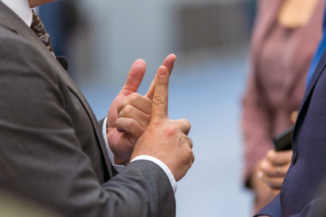 Close-up of a man in a grey suit using his hands to gesture while speaking to two other people, who are also dressed formally. The focus is on the hand movements, indicating communication or explanation, with blurred figures in the background.