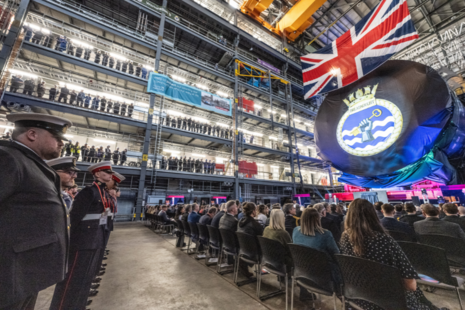 The british flag is hanging from the ceiling of a building, there are people sitting down and a stage.