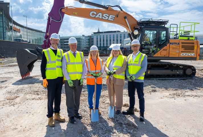 A group of people in helmets and high vis vests stand on bare earth in front of a digger. The two women in the centre each hold a spade.