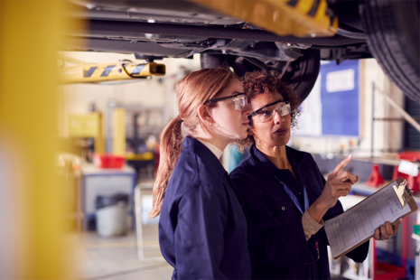 Tutor With Student Looking Underneath Car On Hydraulic Ramp