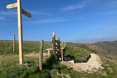 Photo of a section of coastal path showing a gravel track, a stile over wooden fencing and a sign post.
