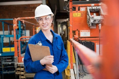 A young female apprentice dressed in blue overalls and a white hard hat at her work place