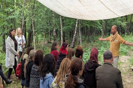 Photograph of a group of people standing under an awning in a forest clearing with the trainer facing them.
