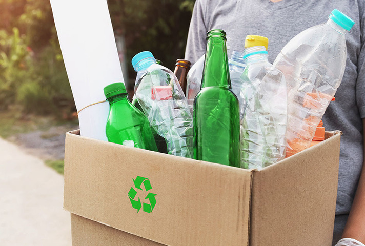 Person holding a cardboard box with a green recycling symbol, filled with green glass bottles and clear plastic bottles, in an outdoor setting with greenery and sunlight.