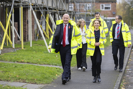 John Healey, Natalie Elphicke-Ross and others visit Service Family Accommodation. They walk along an SFA street in high vis jackets and the houses they pass have scaffolding up. 