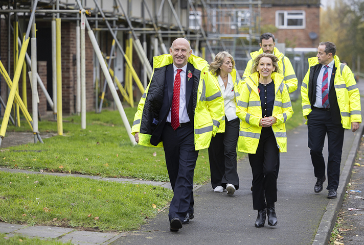 John Healey, Natalie Elphicke-Ross and others visit Service Family Accommodation. They walk along an SFA street in high vis jackets and the houses they pass have scaffolding up.