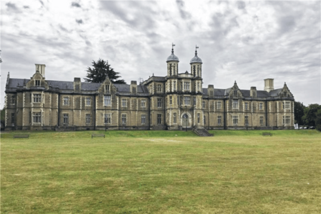 A large historic stone building with twin domed towers, tall sash windows, and ornate details. It stands on a wide grassy lawn under an overcast sky, with several benches in front, conveying a grand yet serene atmosphere.