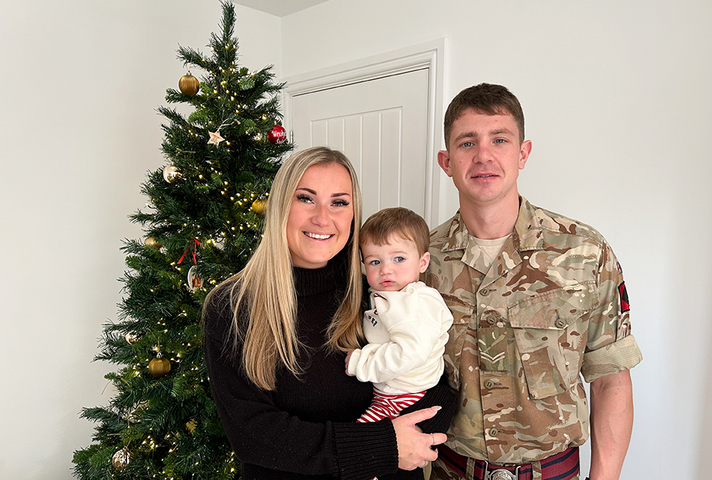 A man in an Army uniform stands next to a woman holding a small child. They stand next to a Christmas tree.