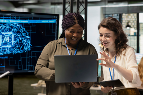 2 people are smiling and looking at a laptop, behind them is a screen with blue lines and AI text.