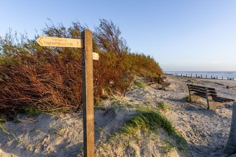Photograph of a signpost about the coast path, set in a sand dune with the sea in the background.