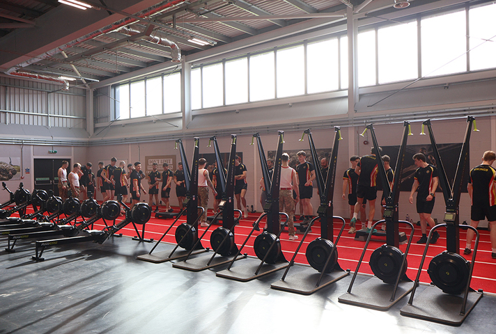 A large group of soldiers stand on one side of a big gym, behind cardio equipment. They are getting ready to train. 