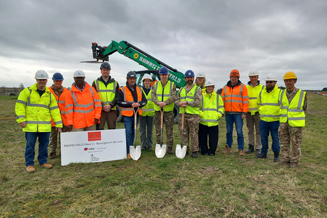 A group of people in high vis stand in a line on an open grassy area. Behind them is a digger. Three of them hold shovels and there is a sign propped against them reading "Prometheus Wave 2 - Bassingbourn Barracks".