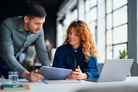 Two people at a desk review a tablet together, with a laptop open beside them in a bright office workspace.