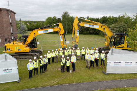 A group of people wearing high visibility jackets and hard hats smile at the camera. They stand in front of two yellow construction vehicles.