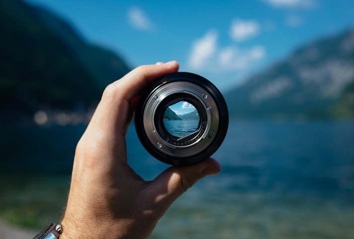 Hand holds camera lens focusing on a lake. (Credit: Unsplash)