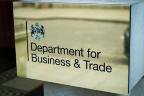 Metal plaque mounted on a building wall displaying the UK government crest and the text “Department for Business & Trade†in black lettering on a brushed metallic background.