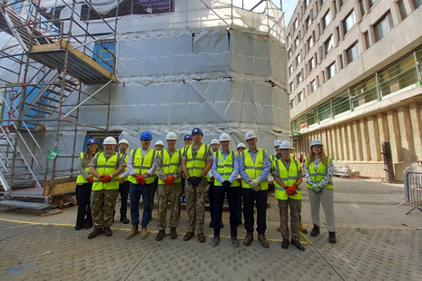 A group of people stand in front of a building covered in scaffolding and white sheets. They all wear helmets and high vis vests.