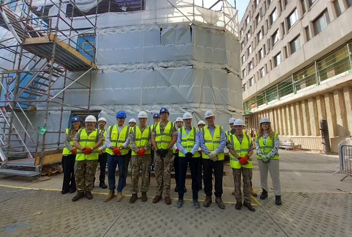 A group of people stand in front of a building covered in scaffolding and white sheets. They all wear helmets and high vis vests.