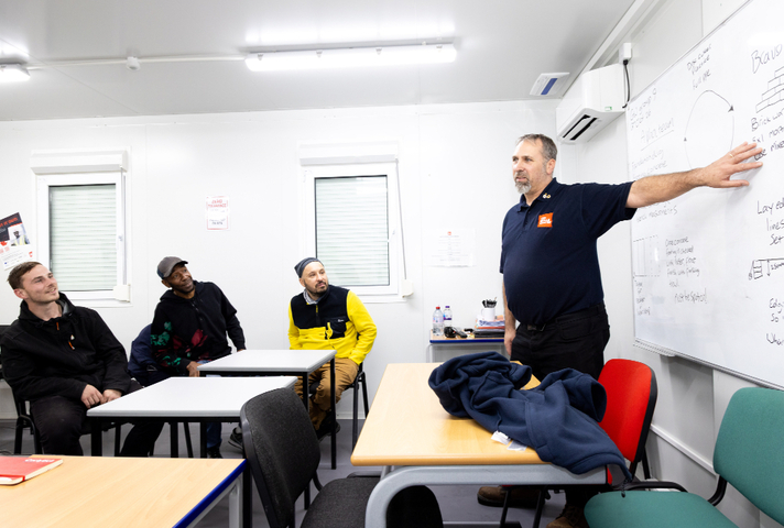 A man in a classroom gestures at notes on a whiteboard while three other men listen to him. 