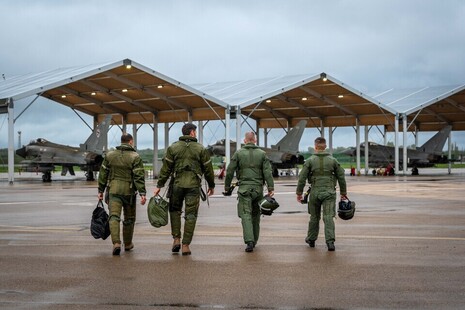 Four personnel in uniform walk towards stationary fighter jets